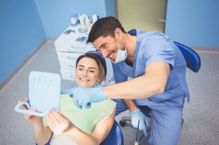 Dentist shows patient the results of treatment with a mirror, examinating teeth with dental equipment in dental office. Young handsome male doctor in medical facial mask and smiling happy woman.の写真素材