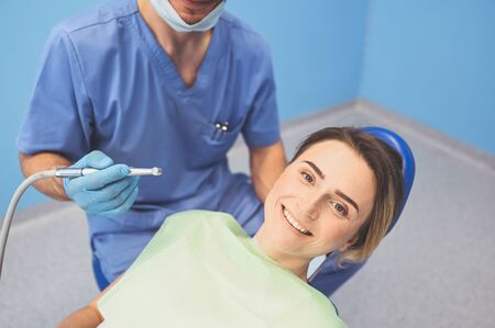 Dentist examining a patients teeth using dental equipment in dentistry office. Stomatology and health concept. Young handsome male doctor in disposable medical facial mask, smiling happy woman.の写真素材