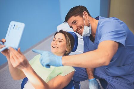 Dentist shows patient the results of treatment with a mirror, examinating teeth with dental equipment in dental office. Young handsome male doctor in medical facial mask and smiling happy woman.の写真素材