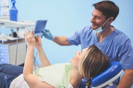 Dentist shows patient the results of treatment with a mirror, examinating teeth with dental equipment in dental office. Young handsome male doctor in medical facial mask and smiling happy woman.の写真素材