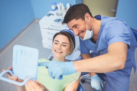 Dentist shows patient the results of treatment with a mirror, examinating teeth with dental equipment in dental office. Young handsome male doctor in medical facial mask and smiling happy woman.の写真素材