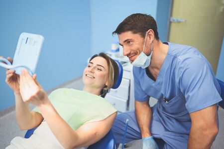 Dentist shows patient the results of treatment with a mirror, examinating teeth with dental equipment in dental office. Young handsome male doctor in medical facial mask and smiling happy woman.の写真素材