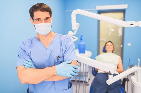 Young smiling handsome male doctor in medical uniform, disposable medical facial mask with equipment in dentistry office and happy patient in the dental chair. Stomatology concept. Copy spaceの写真素材