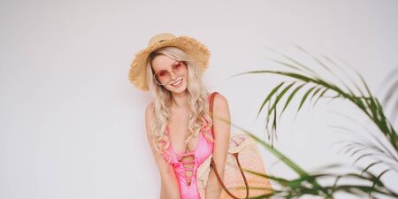 Young sexy smiling happy blonde in a pink swimsuit, a straw hat, sunglasses, excited to present product. Woman on a white background with palm green leaves. Summer sales concept copy space mock upの写真素材