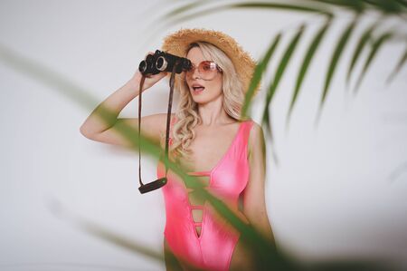 Young sexy smiling happy blonde in a pink swimsuit, a straw hat, sunglasses, excited to present product. Woman on a white background with palm green leaves. Summer sales concept copy space mock upの写真素材