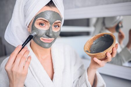 Cosmetology, skin care, face treatment, spa, natural beauty concept. Beautiful smiling woman at home in bathrobe with a towel applying face clay mask against acne to rejuvenate problem skinの写真素材