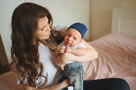 Close up portrait of happy young mom holding her crying baby in blue jeans and white t-shirt and cap. Young happy family, mother playing with cute emotional little newborn child son in the bedroom.の写真素材
