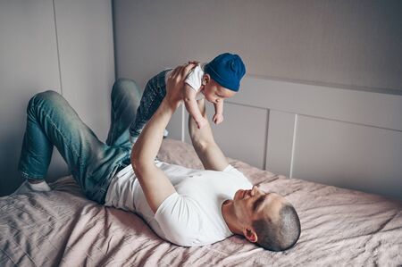 Close up portrait of happy young dad father holding his baby in blue jeans and white t-shirt and cap. Young happy family, dad playing with cute emotional little newborn child son in the bedroom.の写真素材