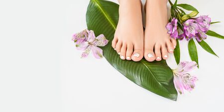 Beautiful perfect female skin legs feet top view with tropical flowers and green palm leaf. Nail polish, care and clean, spa pedicure treatment in white. Concept on a white background. Copy spaceの写真素材