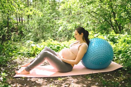 Beautiful young pregnant woman doing exercising with fitness pilates blue ball in park outdoor. Sitting and relaxing on pink yoga mat. Active future mother sport lifestyle. Healthy pregnancy concept.の写真素材