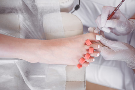 Pedicurist doing white nail polish on client legs using shellac lamp. Professional medical pedicure procedure. Foot treatment in SPA salon. Podiatry clinic. Beautician doctor hands in white glovesの写真素材