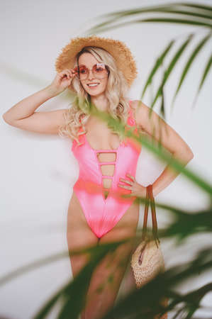 Young smiling happy blonde in a pink swimsuit, a straw hat, sunglasses, excited to present product. Woman on a white background with palm green leaves. Summer sales concept copy space mock upの写真素材