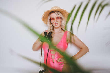 Young smiling happy blonde in a pink swimsuit, a straw hat, sunglasses, excited to present product. Woman on a white background with palm green leaves. Summer sales concept copy space mock upの写真素材