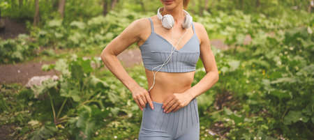 Young happy beautiful slim fit girl in blue sportswear with big white headphones doing squats exercise with fitness training gum in the summer green park outdoor. Girl with perfect body, sport yogaの写真素材