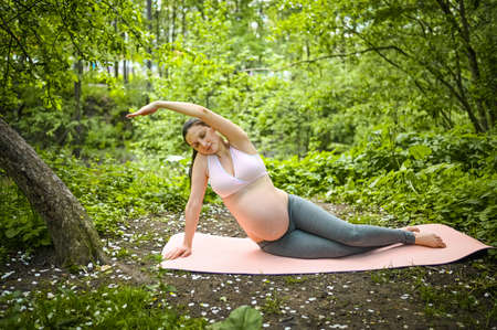Beautiful young pregnant woman doing yoga exercising in park outdoor. Sitting and relaxing on pink yoga mat. Active future mother sport lifestyle. Healthy pregnancy conceptの写真素材