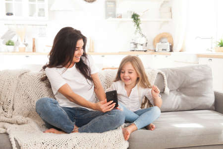 Attractive young mom and little daughter playing online games at home using smartphone, having video chat or watching movie. Distance education. Video conference or virtual event on quarantineの写真素材