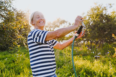 Happy elderly senior woman having fun watering plants with hose in summer garden. Drops of water in backlight. Farming, gardening, agriculture, old age people. Growing organic vegetables on farm.の写真素材