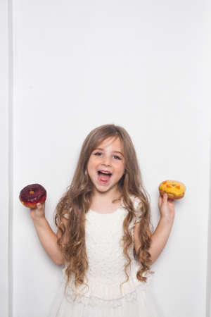 Little happy cute preschool girl is eating colorful donuts on white background isolated. Emotional child is having fun with sweets. Tasty sweet food for kids. Free copy space for text mockupの写真素材