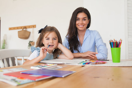 Smiling mother and daughter preparing for lessons and draws at the table with pencils and paints....