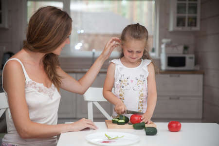 Happy family mother and child posing at home. Beautiful young mom and little daughter having fun and preparing vegetables for salad in a white kitchen in a Scandinavian style interior. Healthy foodの写真素材