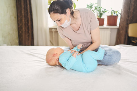 Beautiful mother in protective face mask holding her little cute newborn son protecting him from...