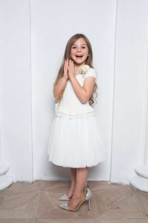 Little princess - excited emotional cute girl in fashion white dress having fun and wearing big mothers sparkle high heels shoes on white background. Free space for text mockupの写真素材