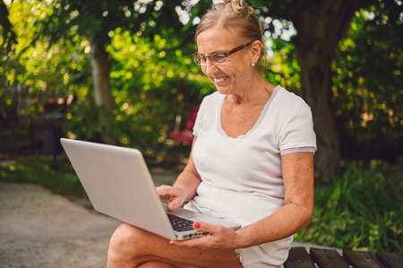 Technology, old age people concept - elderly happy senior old woman working online with laptop computer outdoor in the garden. Remote work, distance education.の写真素材