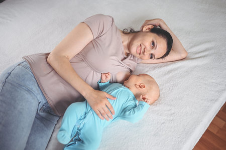 Young mother lies with a newborn cute infant baby in blue jumpsuit on the bed, hugging and breastfeeds him with breast milk. Healthy child, concept of hospital and happy motherhood. Nursery.の写真素材