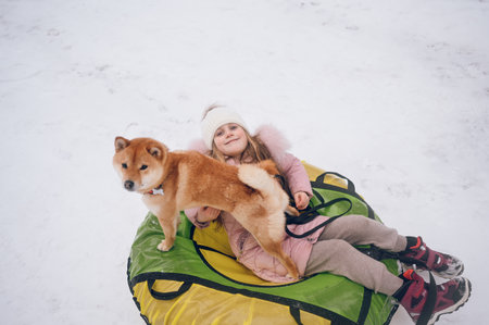 Little cute girl in pink warm outwear having fun with red shiba inu dog rides inflatable snow tube in snowy white cold winter outdoors. Family sport vacation activitiesの写真素材