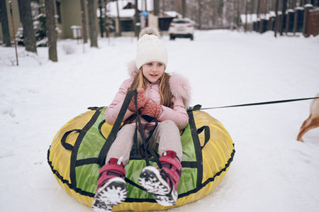 Little cute girl in pink warm outwear having fun rides inflatable snow tube in snowy white cold winter outdoors. Family sport vacation activitiesの写真素材