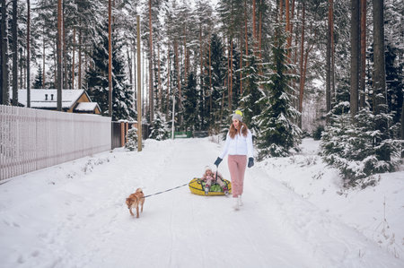 Happy mother and little cute girl in pink warm outwear walking having fun rides inflatable snow tube with red shiba inu dog in snowy white cold winter forest outdoors. Family sport vacation activitiesの写真素材
