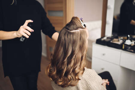Hair stylist prepares woman makes curls hairstyle with curling iron. Long light brown natural hairの写真素材