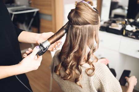 Hair stylist prepares woman makes curls hairstyle with curling iron. Long light brown natural hairの写真素材