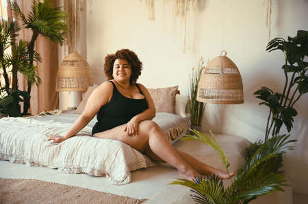 Beautiful curvy oversize African black woman afro hair lying on bed eco friendly cozy bedroom interior design with home plants. Body imperfection, body acceptance, body positive and diversity concept.の写真素材