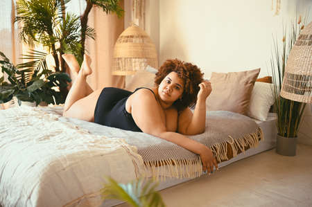 Beautiful curvy oversize African black woman afro hair lying on bed eco friendly cozy bedroom interior design with home plants. Body imperfection, body acceptance, body positive and diversity concept.の写真素材