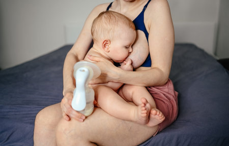 Young mother with newborn cute infant naked baby boy, holding him on arms, using breast pump hugging...