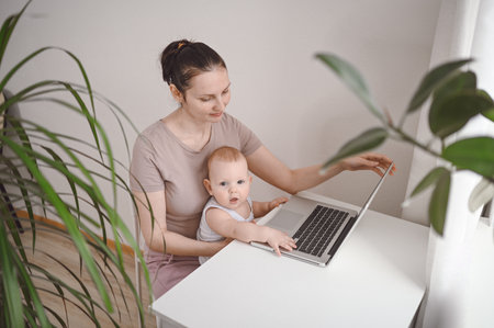 Young mother work studying from home with laptop computer during quarantine, little cute toddler baby on lap. Home office, parenthood. Remote work, distance education. Video conference virtual eventの写真素材
