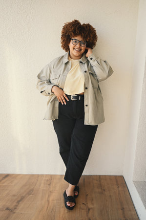 Beautiful happy smiling curvy plus size African black woman afro hair posing in beige t-shirt, jeans, stylish glasses on beige background. Body imperfection, acceptance body positive diversity conceptの写真素材