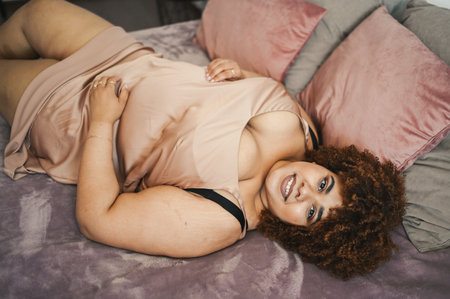 Beautiful curvy plus size African black woman afro hair lying on bed in silk powder pink dress cozy bedroom interior design. Body imperfection, body acceptance, body positive and diversity concept.の写真素材