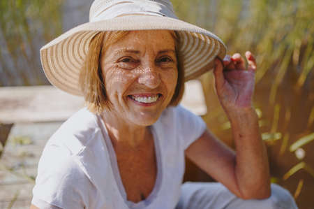 Close up portrait of happy smiling elderly senior woman in straw hat sitting on wooden pier by lake. Farming, gardening, agriculture, retired old age people. Country summer rest.の写真素材