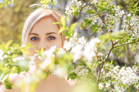Close up young beautiful blonde woman with blue eyes posing in blooming spring garden with white...