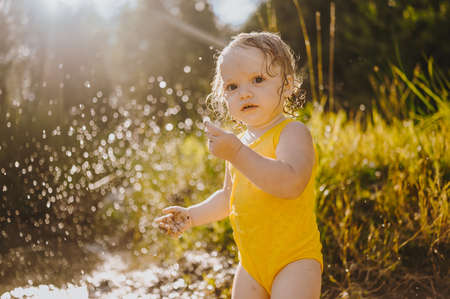 Little funny cute blonde girl child toddler in yellow wet bodysuit playing by the lake waterside shore at sunset outside. Baby with mud on hands and legs. Water activities at summerの写真素材