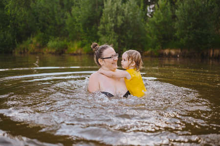 Little funny cute blonde girl child toddler in yellow bodysuit laughing learns swim outside at summer lake. Plus size body positive smiling woman mother baby swimming in natural pool. Water splashesの写真素材