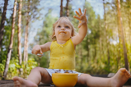 Little funny cute blonde girl child toddler with dirty clothes and face eating baby food fruit or...