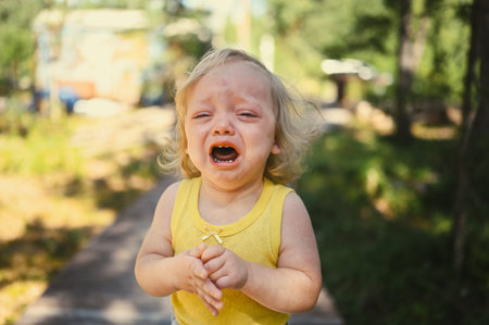 Close up portrait of little funny cute blonde girl child toddler in yellow bodysuit crying outside at summer. Childish tantrum. Healthy childhood conceptの写真素材