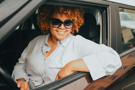 Happy young smiling African American woman red afro haired driver in sun glasses sitting in new brown car, smiling looking at camera enjoying journey. Driving courses and life insurance concept.の写真素材