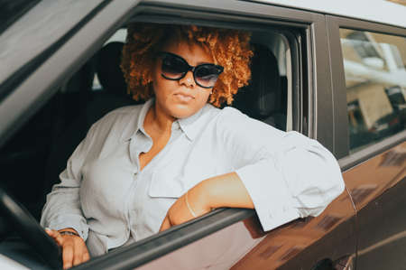 Happy young smiling African American woman red afro haired driver in sun glasses sitting in new brown car, smiling looking at camera enjoying journey. Driving courses and life insurance concept.の写真素材