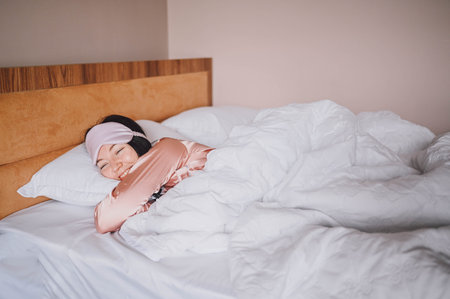 Attractive smiling young woman in pink silk pajamas and eye sleeping mask stretching in bed waking...