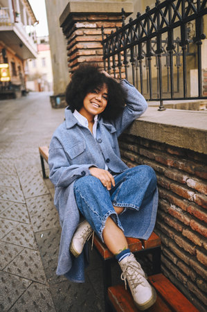 Fashion street style portrait of attractive young natural beauty African American woman with afro hair in blue coat and jeans posing outdoors. Happy tourist laughing walks around the city landscapesの写真素材