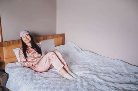 Young happy brunette woman in sleeping mask lying in bed in pink silk pajamas with laptop computer, have online video chat. Blogger or freelancer with remote work at homeの写真素材